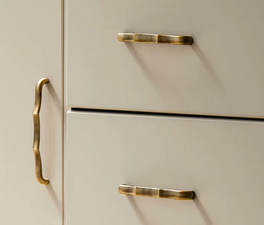 Close-up of cream-colored drawers with polished brass hardware, including a decorative bracket-style handle on the left and two pull handles on the drawers.