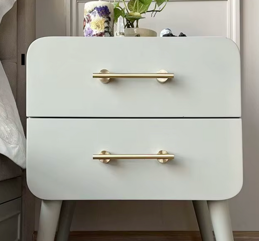 A two-drawer nightstand with a matte white finish and round gold metal handles, shown in a bedroom setting with a vase of flowers on top.