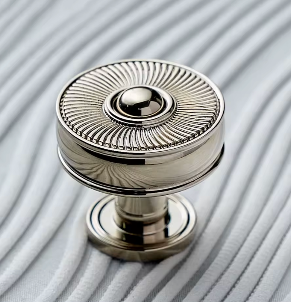 A polished silver cabinet knob with a sunburst ribbed pattern on the top, shown on a textured gray background.