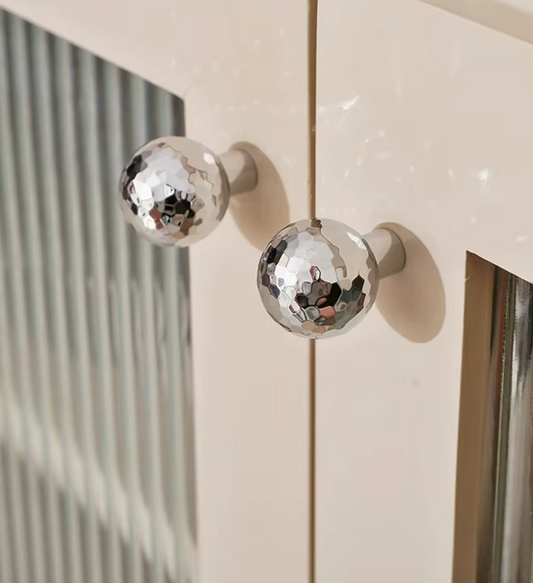 Two round hammered-finish metallic cabinet knobs mounted on cream-colored doors, with a softly blurred interior background visible through the left door.