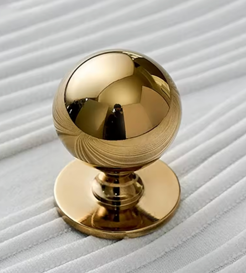 A polished gold spherical cabinet knob with a circular base, shown on a white ribbed fabric background.