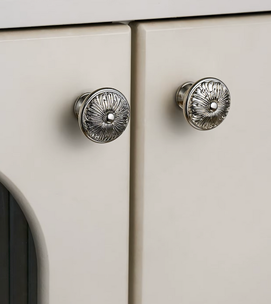 Two round silver cabinet knobs with an embossed floral pattern, shown on cream-colored doors.