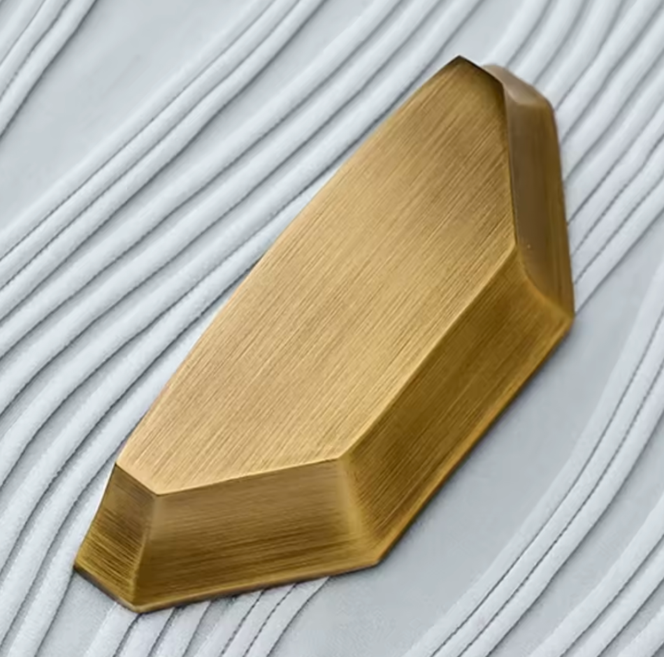 A brushed brass geometric metal block with a faceted, elongated shape, resting on a white textured surface with parallel grooves.