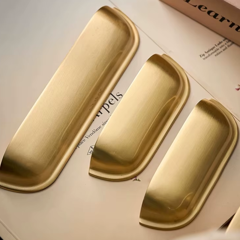 Three elongated brushed-metal drawer pulls in a warm gold finish, arranged diagonally on a light beige surface with printed text underneath.