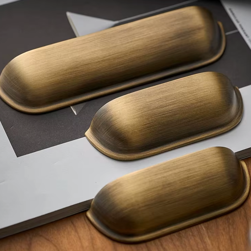 Three elongated oval cabinet knobs in a brushed antique brass finish, arranged diagonally on a styled flat-lay background of dark gray, white, and wood grain.