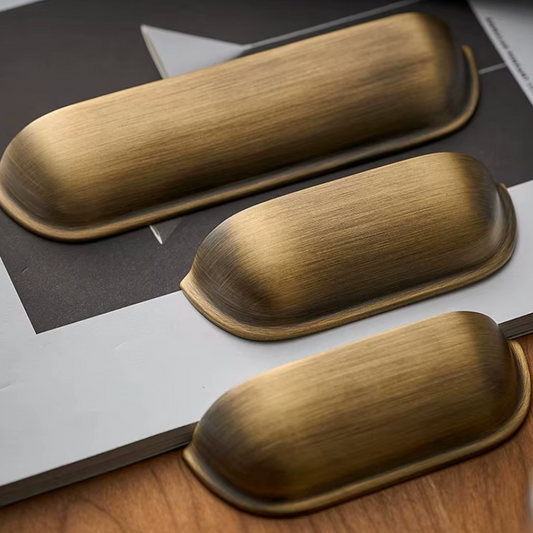 Three elongated oval cabinet knobs in a brushed antique brass finish, arranged diagonally on a styled flat-lay background of dark gray, white, and wood grain.