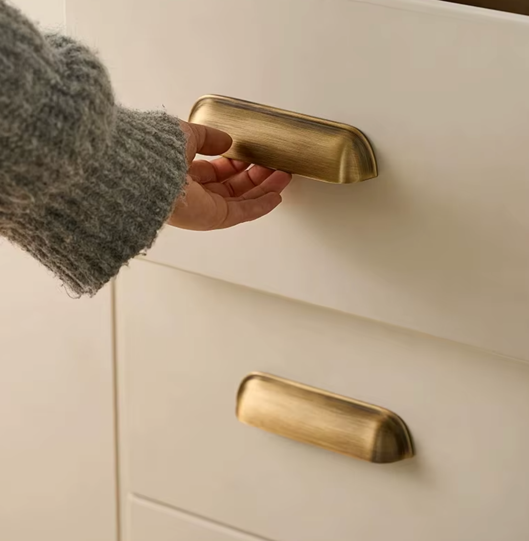 A close-up of a hand in a gray knit sweater pulling a drawer with a brushed brass pull handle, with another matching handle visible on the lower drawer.