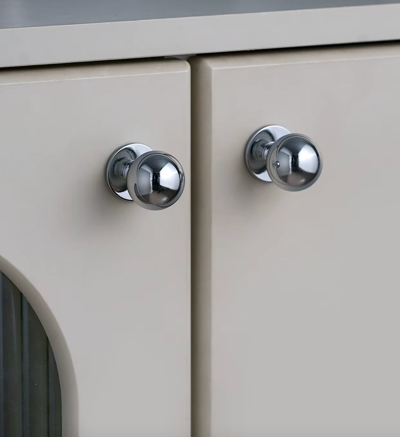 Two round polished chrome cabinet knobs mounted on light beige cabinet doors.