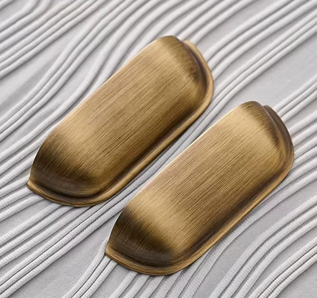 Two oval cabinet knobs with a brushed antique gold finish, displayed on a textured white background with diagonal grooves.