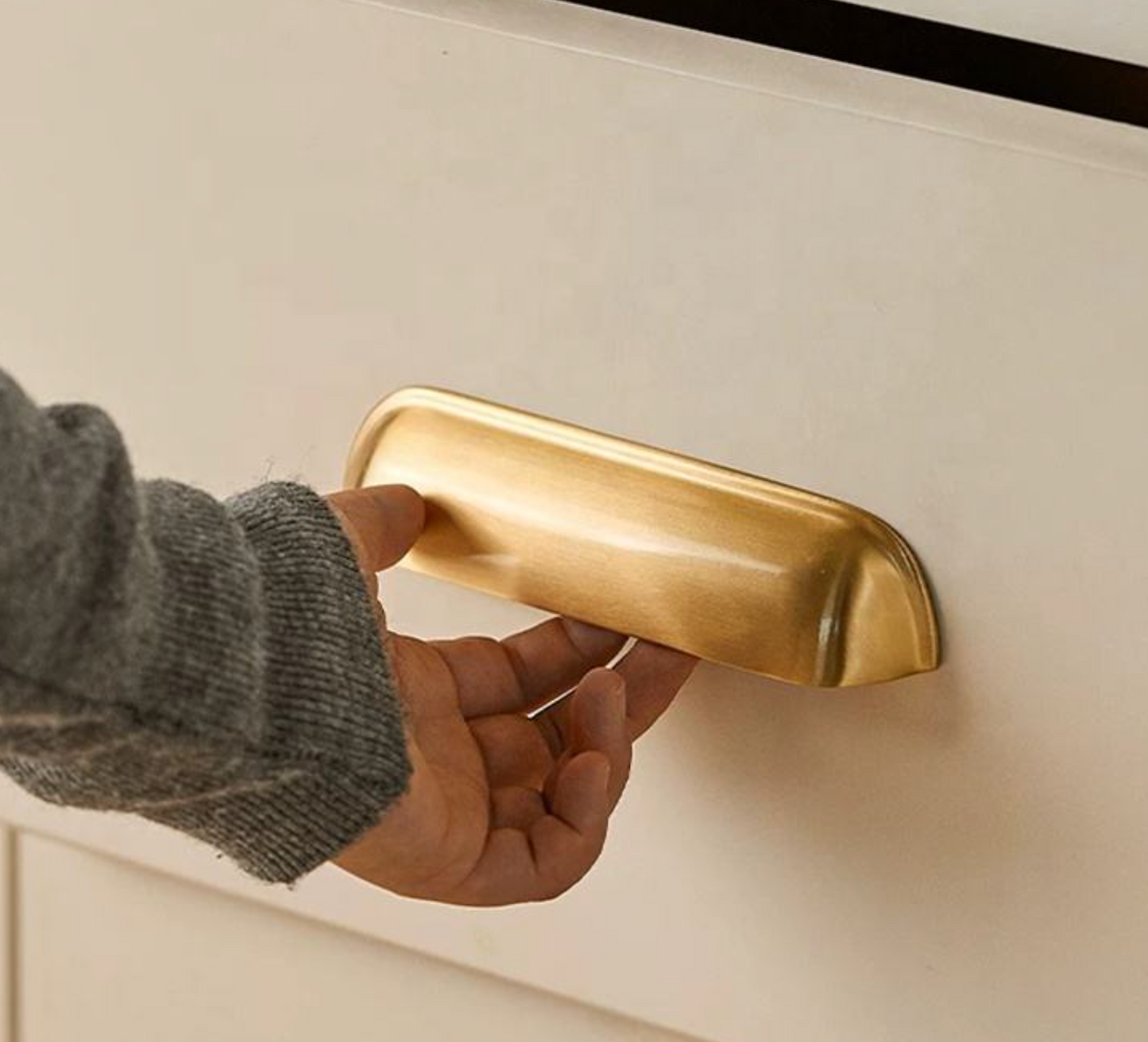 A hand wearing a gray knit sleeve is holding a round, brushed gold metal drawer pull handle mounted on a white drawer front.