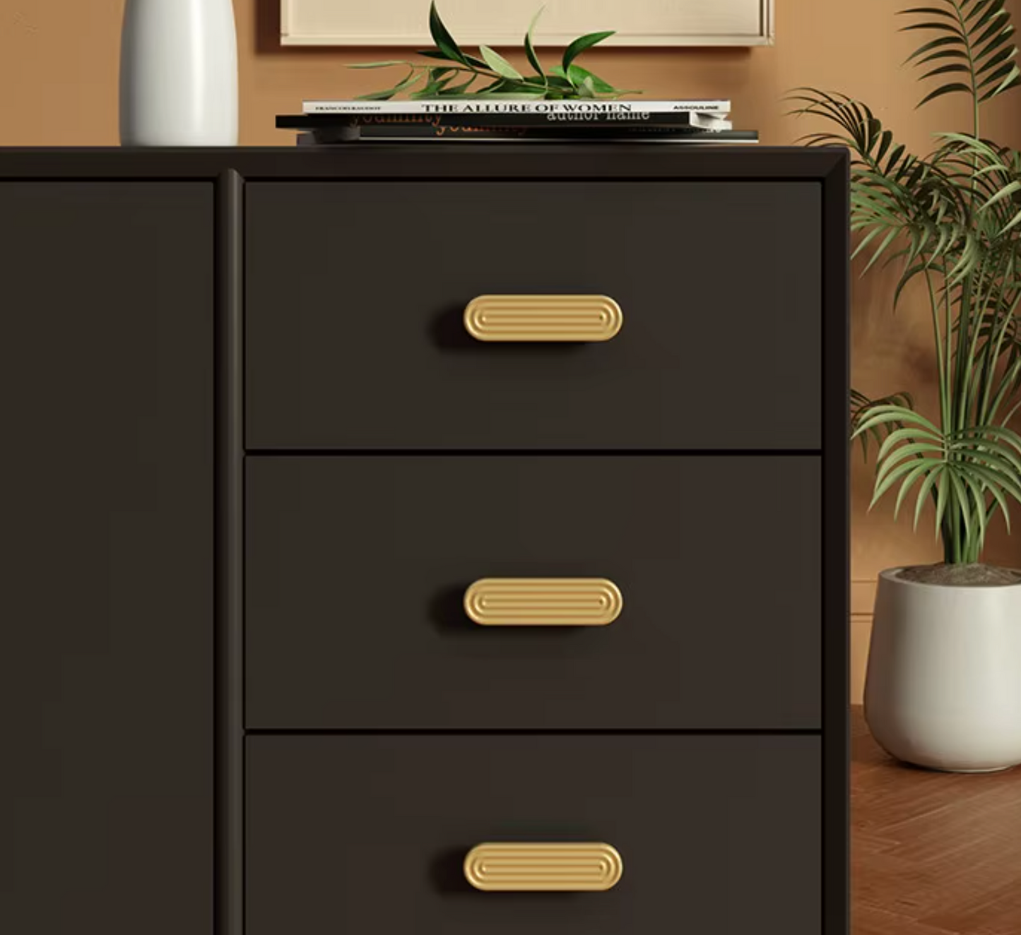 A close-up of a dark brown three-drawer dresser with gold oval pull handles, styled in a room setting with a plant and books on top.
