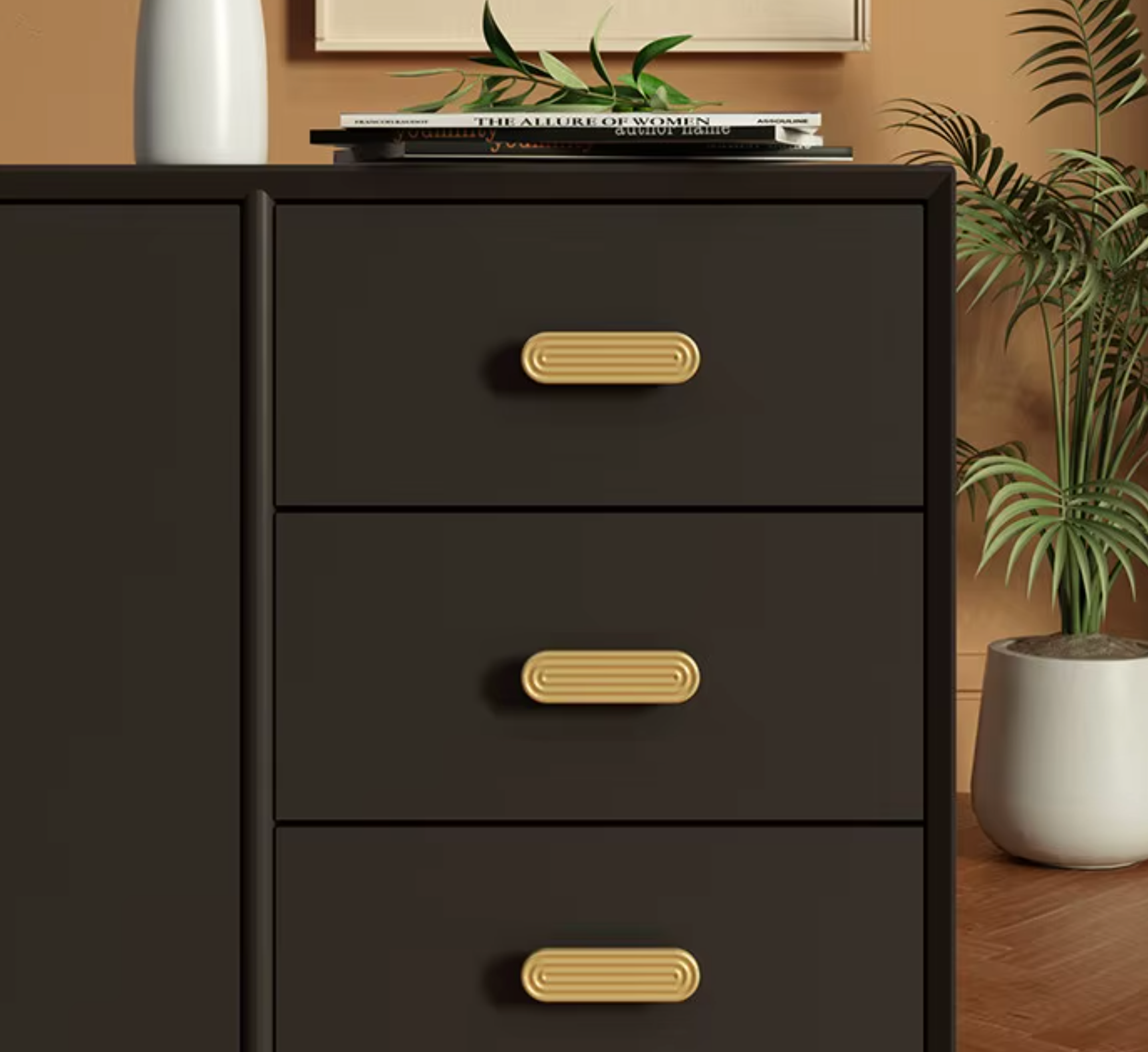 A close-up of a dark brown three-drawer dresser with gold oval pull handles, styled in a room setting with a plant and books on top.