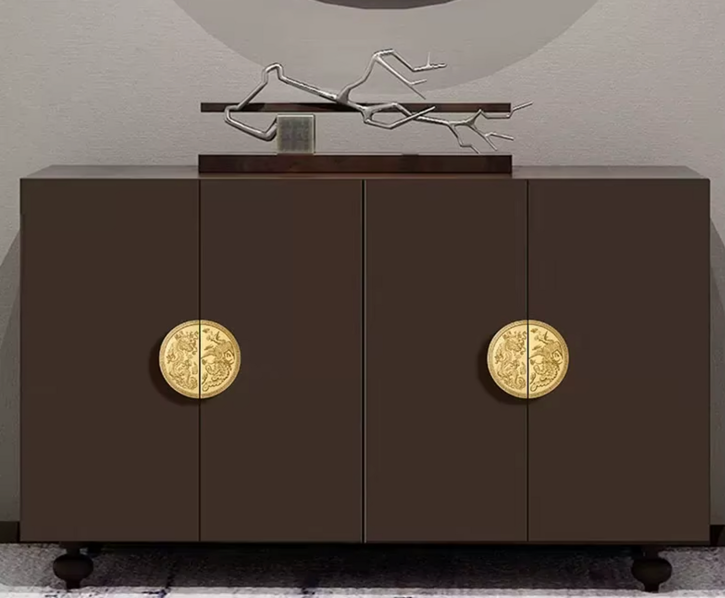 A modern dark brown sideboard cabinet with two doors, each featuring a large circular gold-toned decorative handle, shown against a neutral gray wall with a decorative metal sculpture on top.