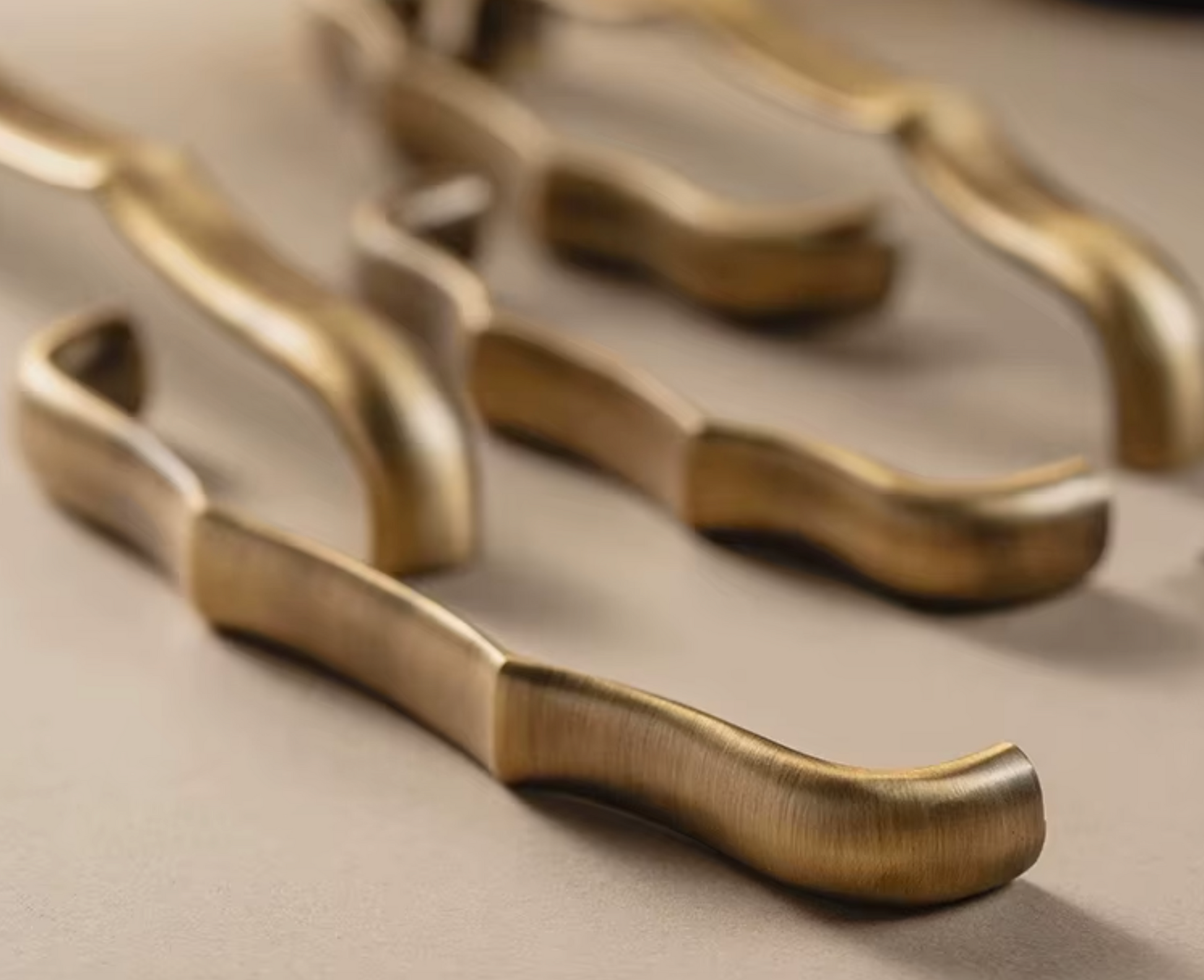A close-up, shallow depth-of-field shot of several brushed brass or bronze kitchen utensils with curved, ergonomic handles resting on a neutral beige surface.