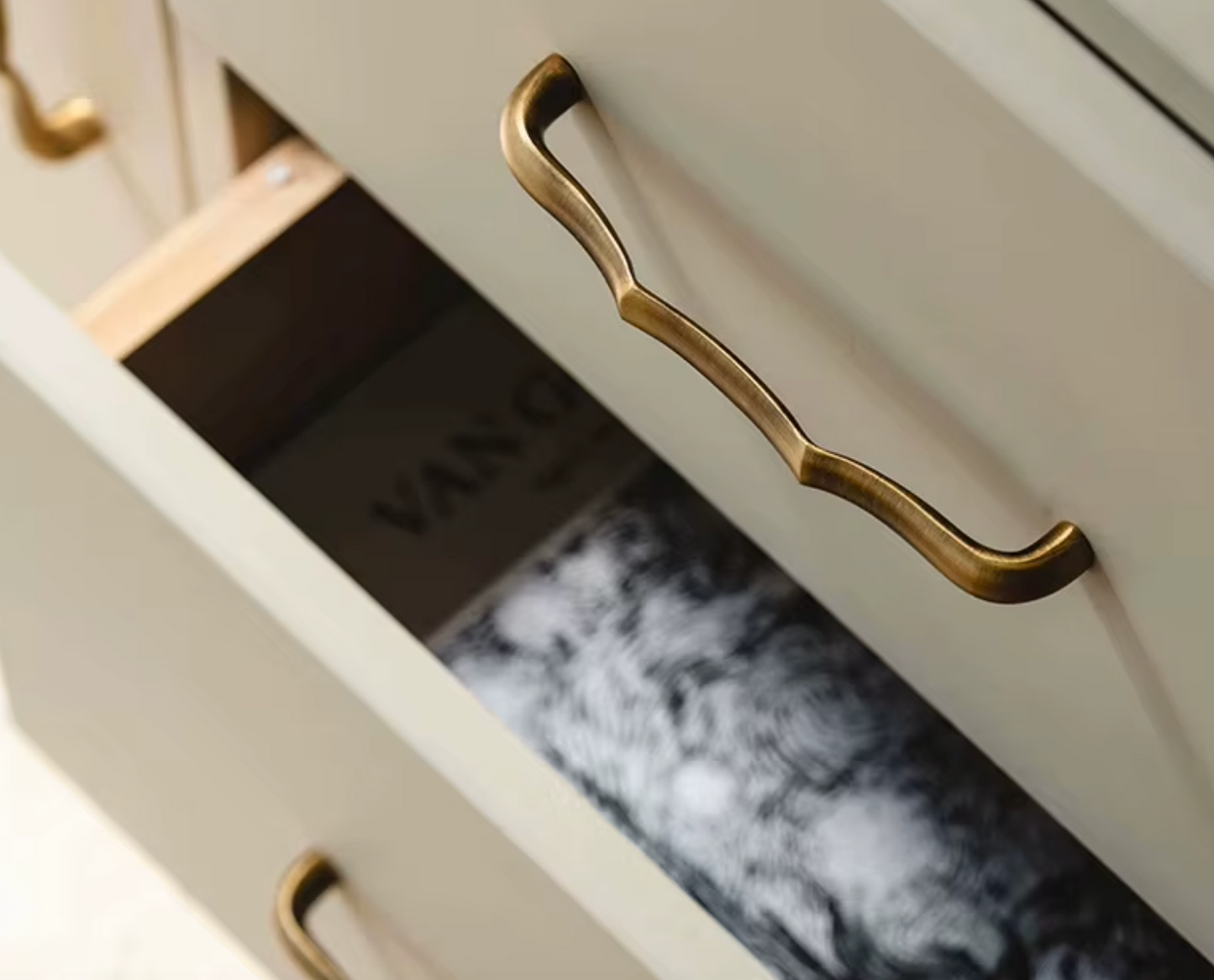 A close-up of a cream-colored wooden drawer featuring a distinctive curved, scalloped antique brass pull handle, with the drawer pulled open to reveal contents.