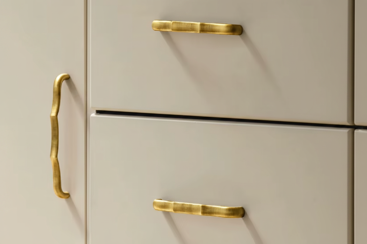 Close-up of cream-colored drawers with polished brass hardware, including a decorative bracket-style handle on the left and two pull handles on the drawers.