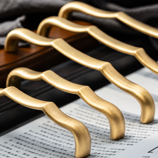 Four curved metallic cabinet handles in a brushed gold finish, shown resting on a dark wooden surface with a dark cloth and printed paper in the background.