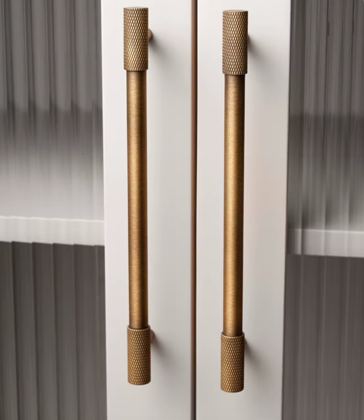 Two long cylindrical cabinet handles in a brushed brass or bronze finish, mounted on white double doors with a blurred gray interior background.
