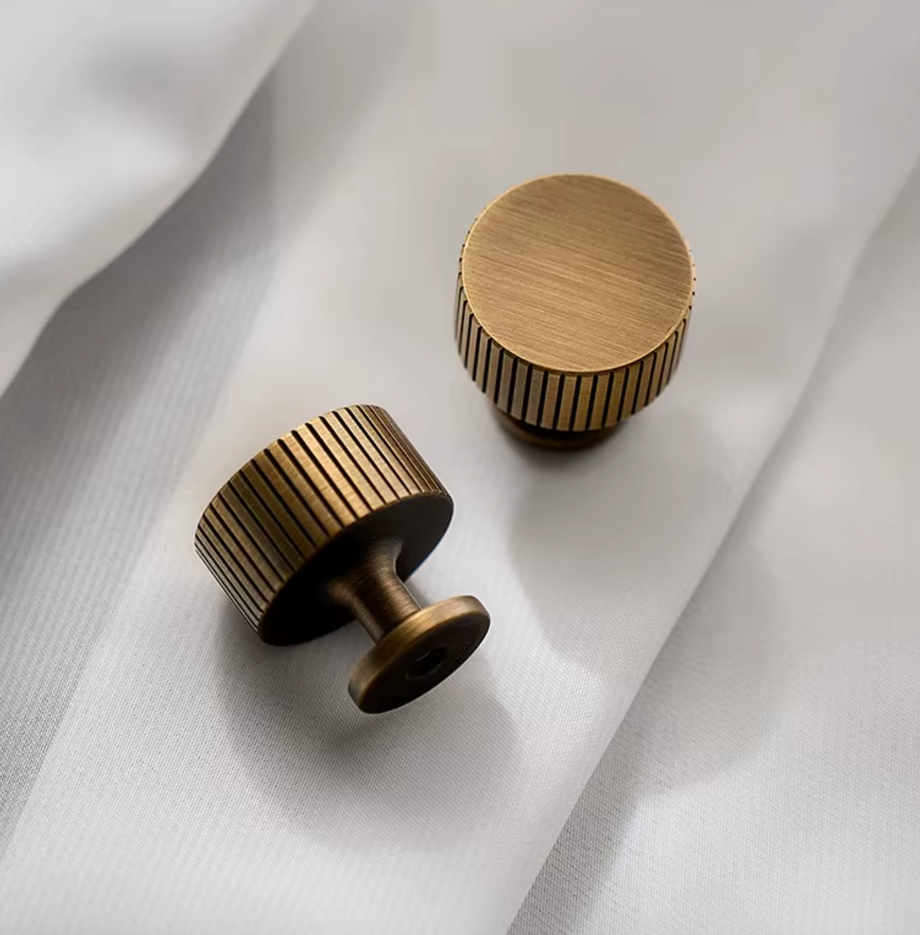 Two round cabinet knobs in an antique brass finish, one showing the side with vertical grooves and the other showing the top and front, resting on a draped white fabric background.