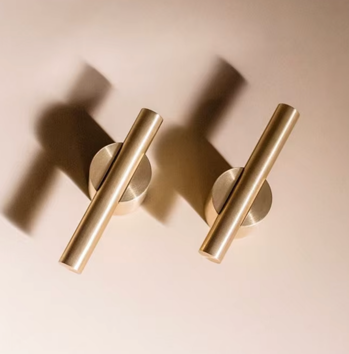 Two modern cylindrical wall-mounted cabinet pulls in a brushed brass or gold finish, shown on a warm beige background with soft shadows.