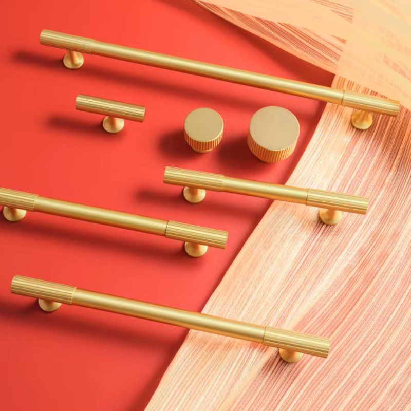 A set of modern brass cabinet hardware including long bar pulls, a shorter horizontal bar pull, two round knobs, and two round pull ends, arranged on a split red and beige background.