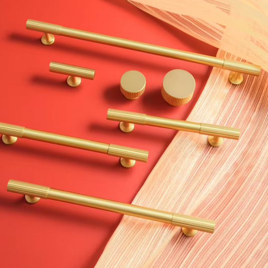 A set of modern brass cabinet hardware including long bar pulls, a shorter horizontal bar pull, two round knobs, and two round pull ends, arranged on a split red and beige background.
