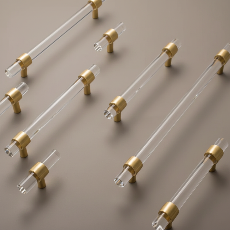 Multiple modern cabinet pulls with clear glass rods and brushed gold metal end caps arranged diagonally on a light gray background.