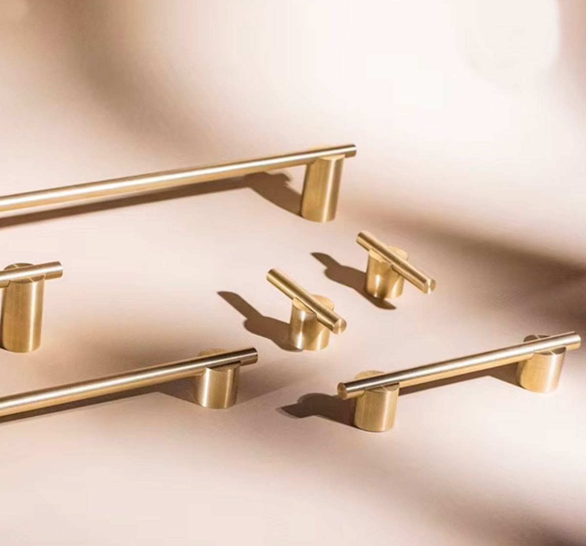 A set of modern linear cabinet pulls in a brushed gold finish, arranged on a warm beige surface with soft, artistic shadows.