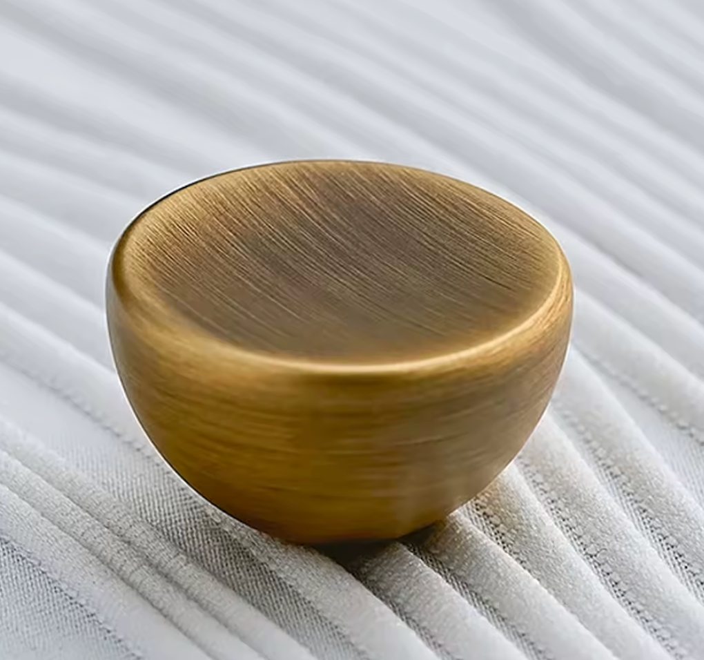 A round, dome-shaped cabinet knob with a brushed brass finish, resting on a white ribbed fabric surface.
