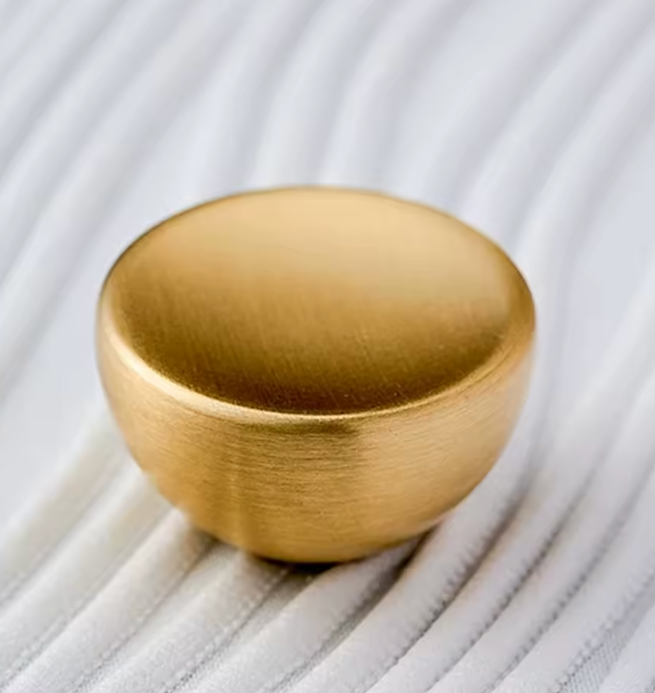 A round, dome-shaped gold cabinet knob with a brushed metal finish, resting on a white ribbed fabric surface.