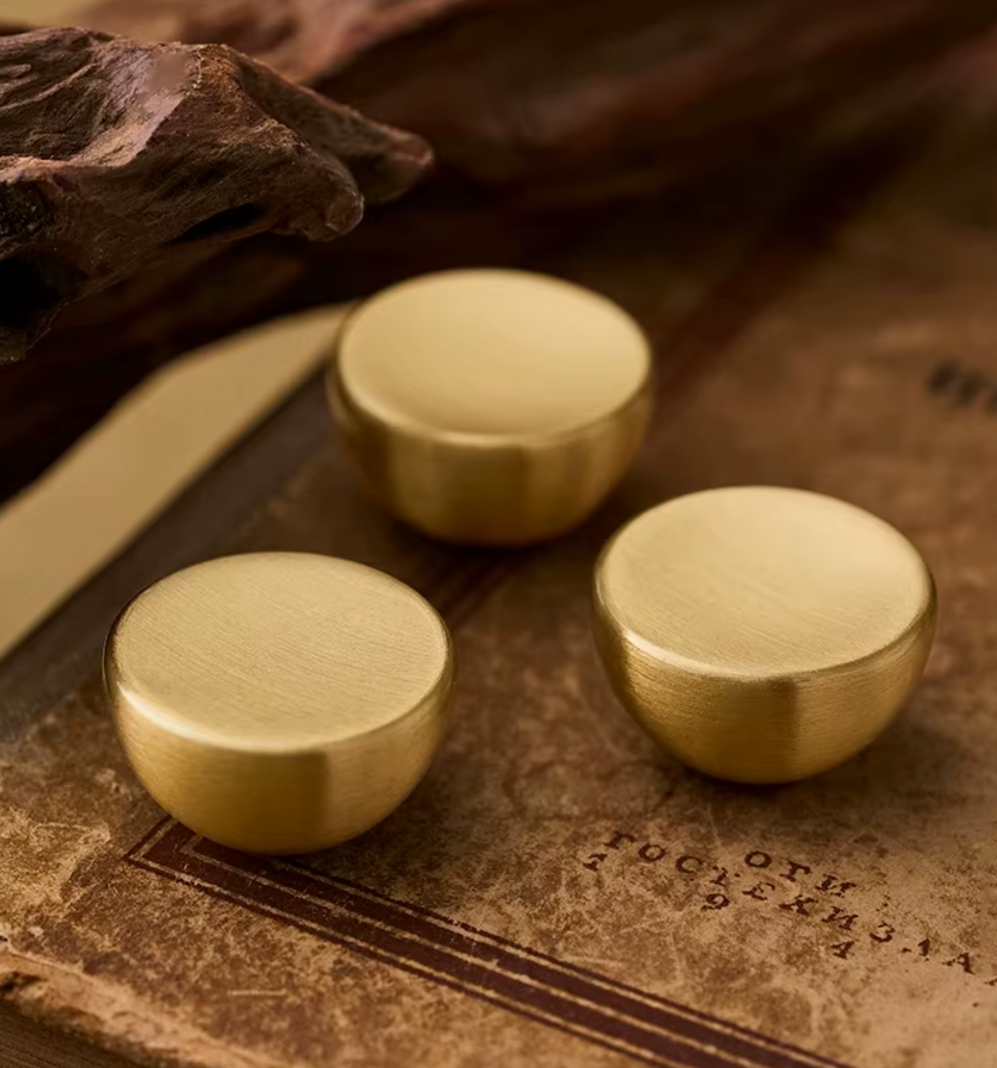 Three round brushed brass or gold-toned metal knobs with a matte finish, arranged on an aged brown book page with a piece of driftwood in the background.