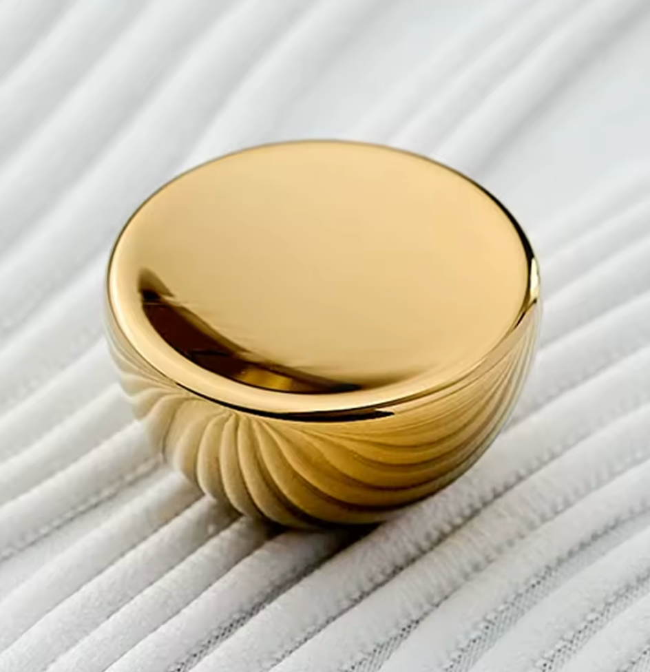 A polished gold metallic knob with a smooth, reflective circular top and a ribbed textured base, resting on a white ribbed fabric surface.