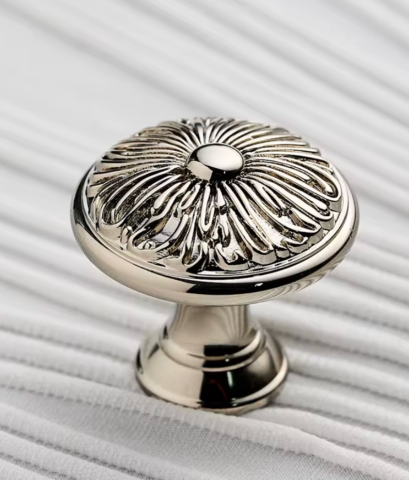 A polished silver cabinet knob with an ornate sunburst or floral relief pattern on the round top, shown on a white ribbed fabric background.