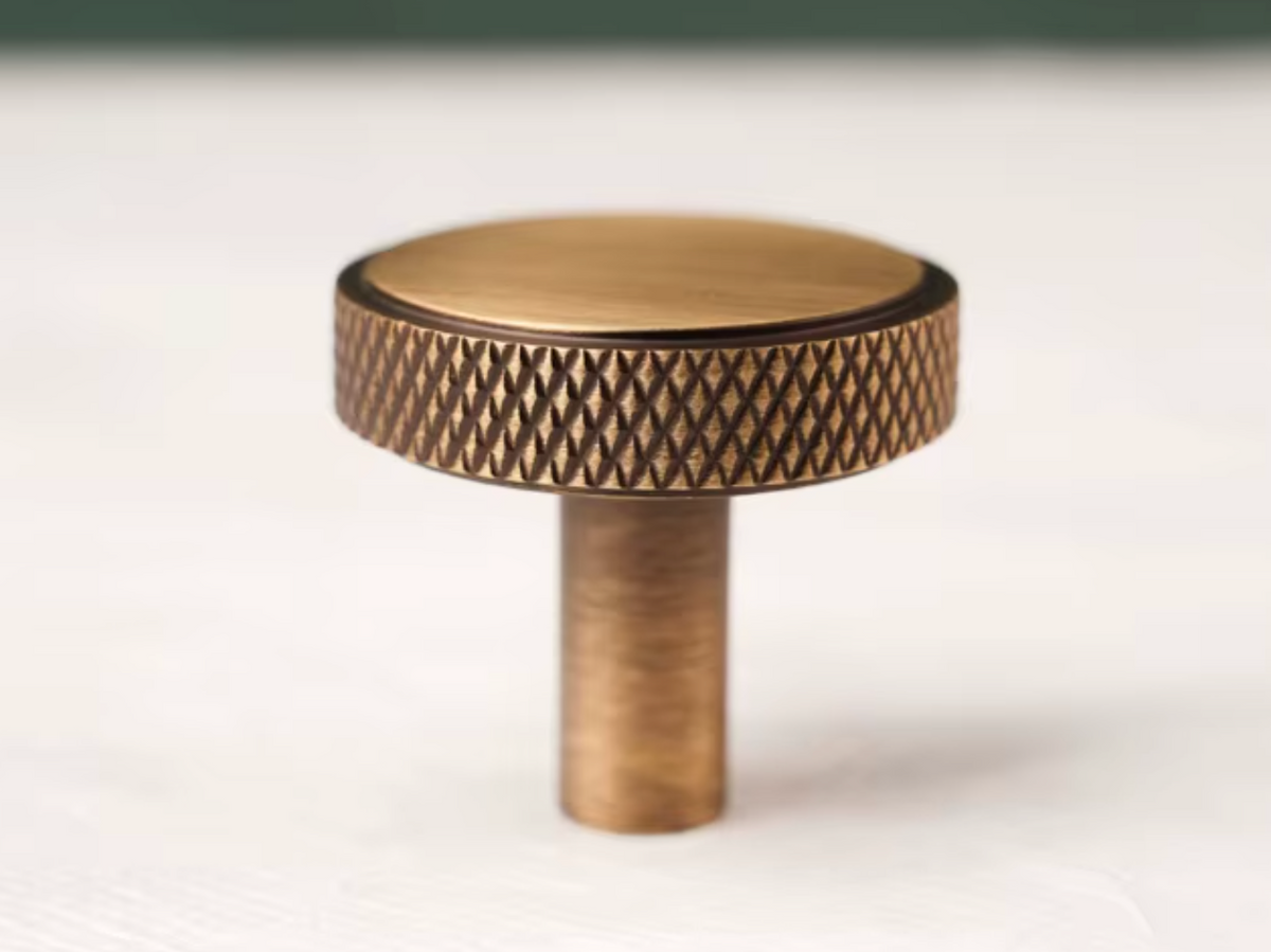 A single round cabinet knob in an antique brass finish, featuring a textured knurled band and a smooth flat top, photographed on a light surface.