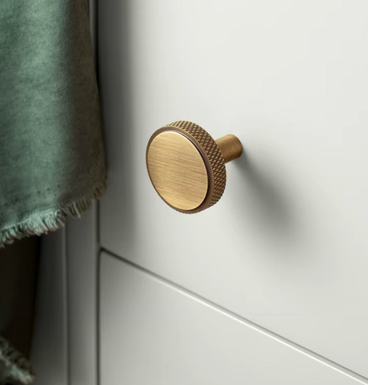 A close-up of a round brushed brass cabinet knob mounted on a white drawer front, with a green textile partially visible on the left.
