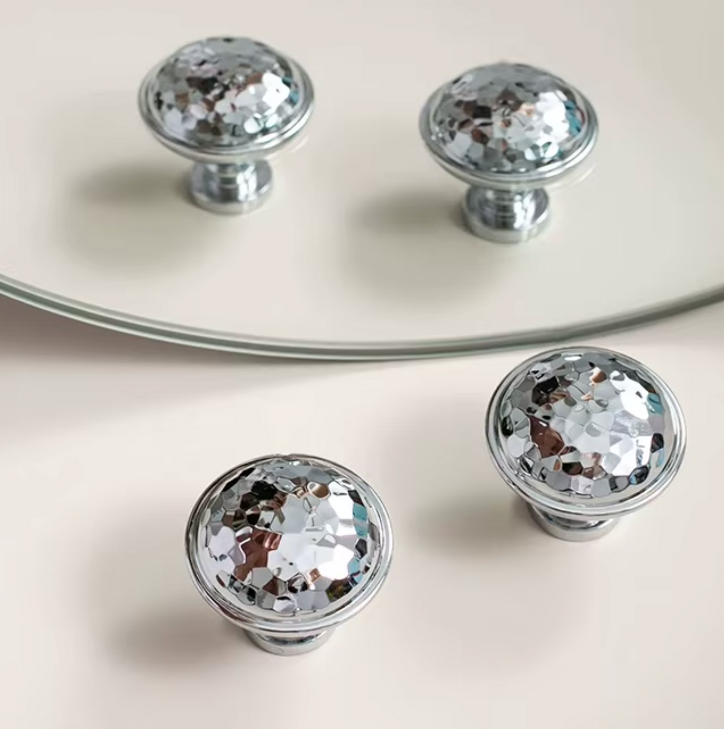 Three round silver cabinet knobs with a faceted, diamond-cut mirrored surface, displayed on a light beige surface with a glass mirror reflecting two of the knobs.
