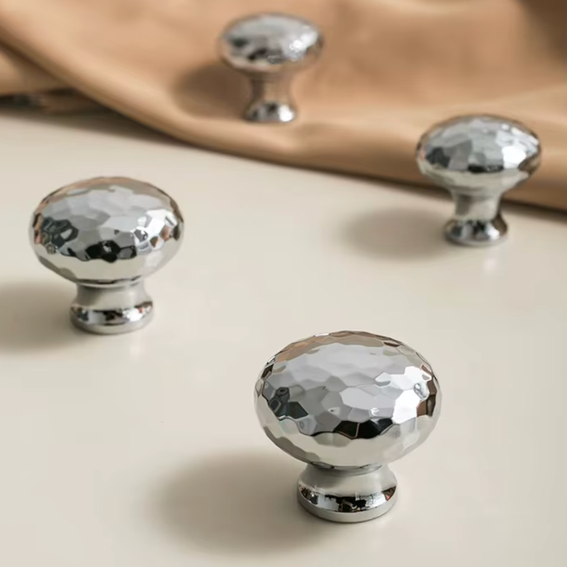 Four shiny silver cabinet knobs with a faceted, hammered finish, displayed on a light beige surface with a soft tan fabric in the background.