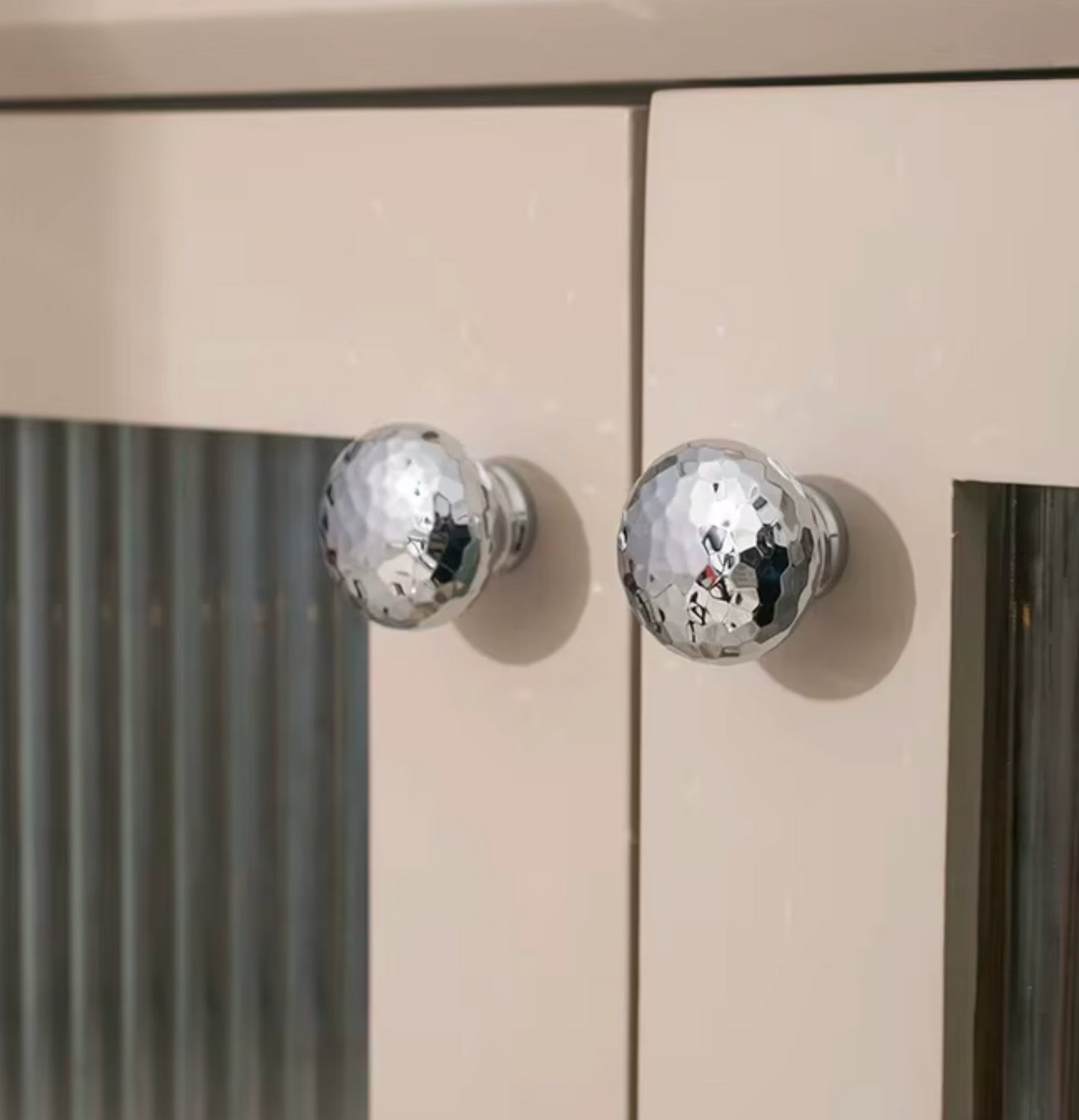 Two round hammered chrome cabinet knobs mounted on a beige cabinet door with a glass panel.