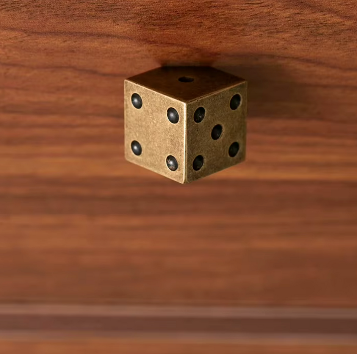 A small brass-colored dice-shaped wall hook or cabinet pull mounted on a warm brown wooden surface.