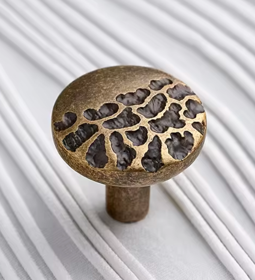 A round brass cabinet knob with a textured, hammered surface and organic cutout patterns, resting on a white ribbed surface.