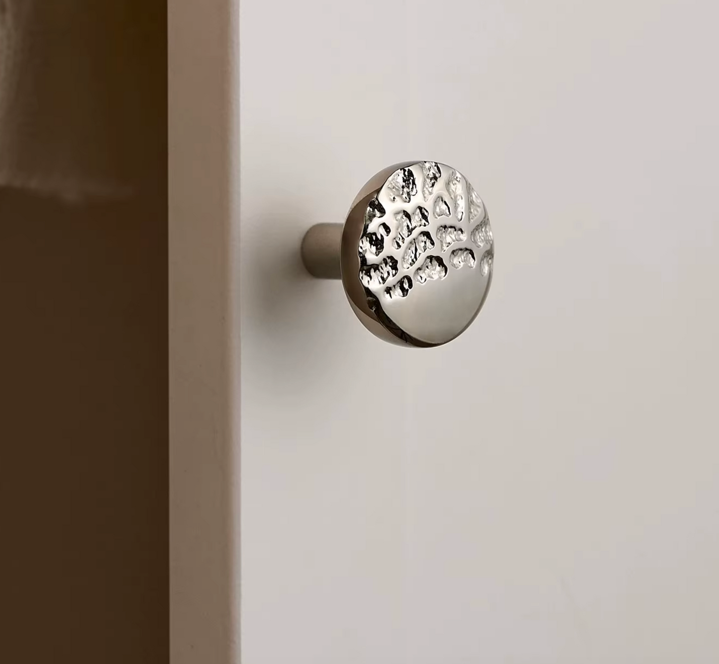 A round silver metal cabinet knob with a textured, hammered pattern on its face, mounted on a white door next to a dark brown frame.