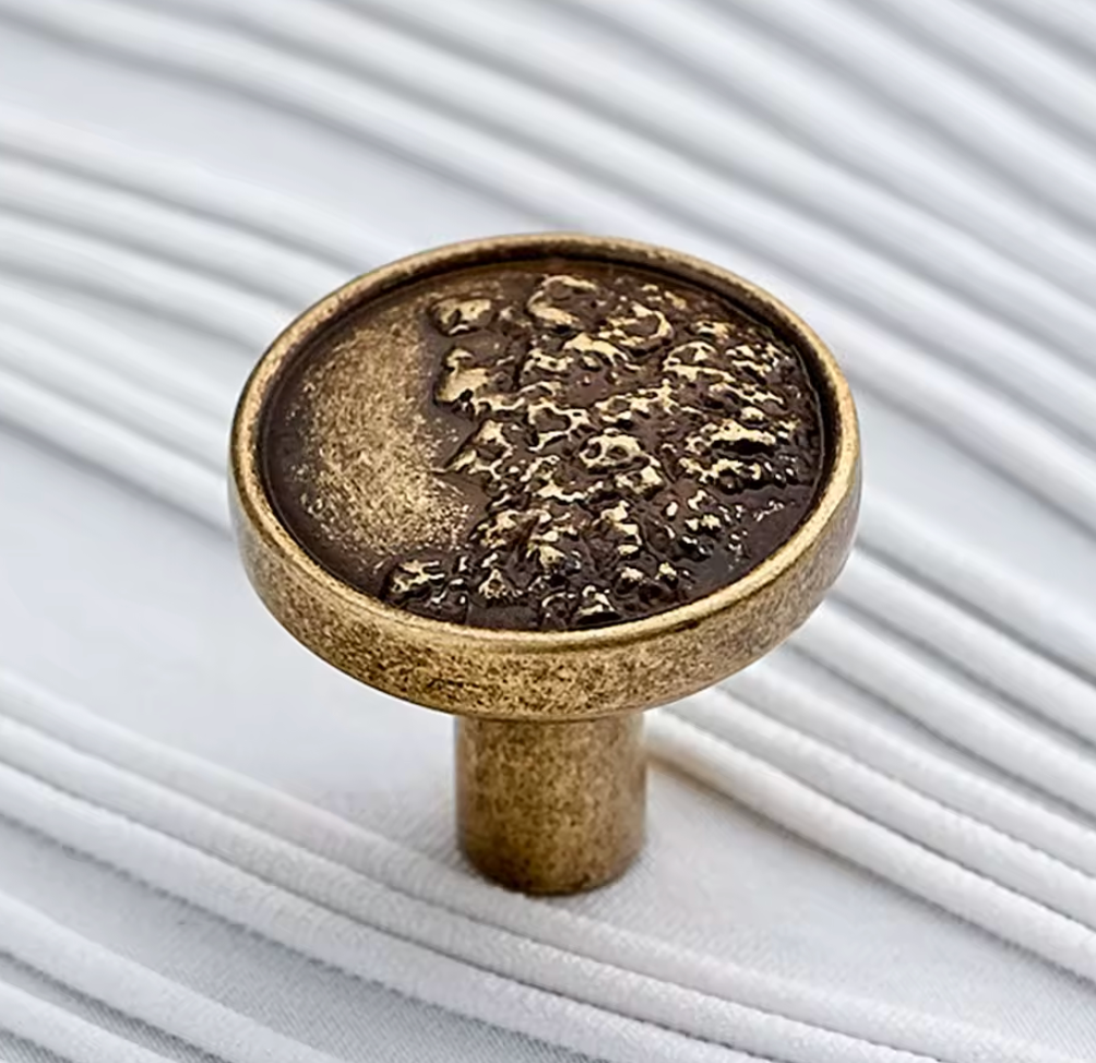 An antique bronze cabinet knob with a textured, distressed finish, shown in a close-up shot against a background of white corded fabric.
