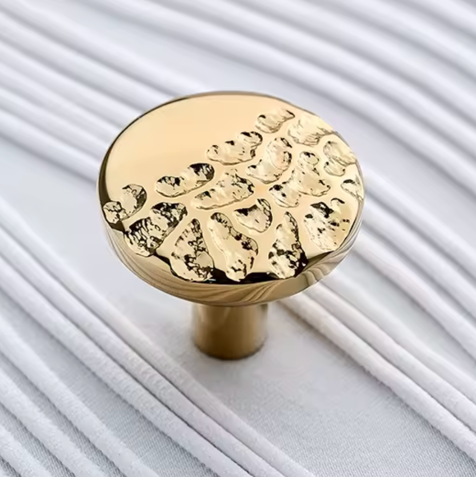 A round gold cabinet knob with a textured, hammered leaf-like pattern on its top surface, resting on a white ribbed fabric background.