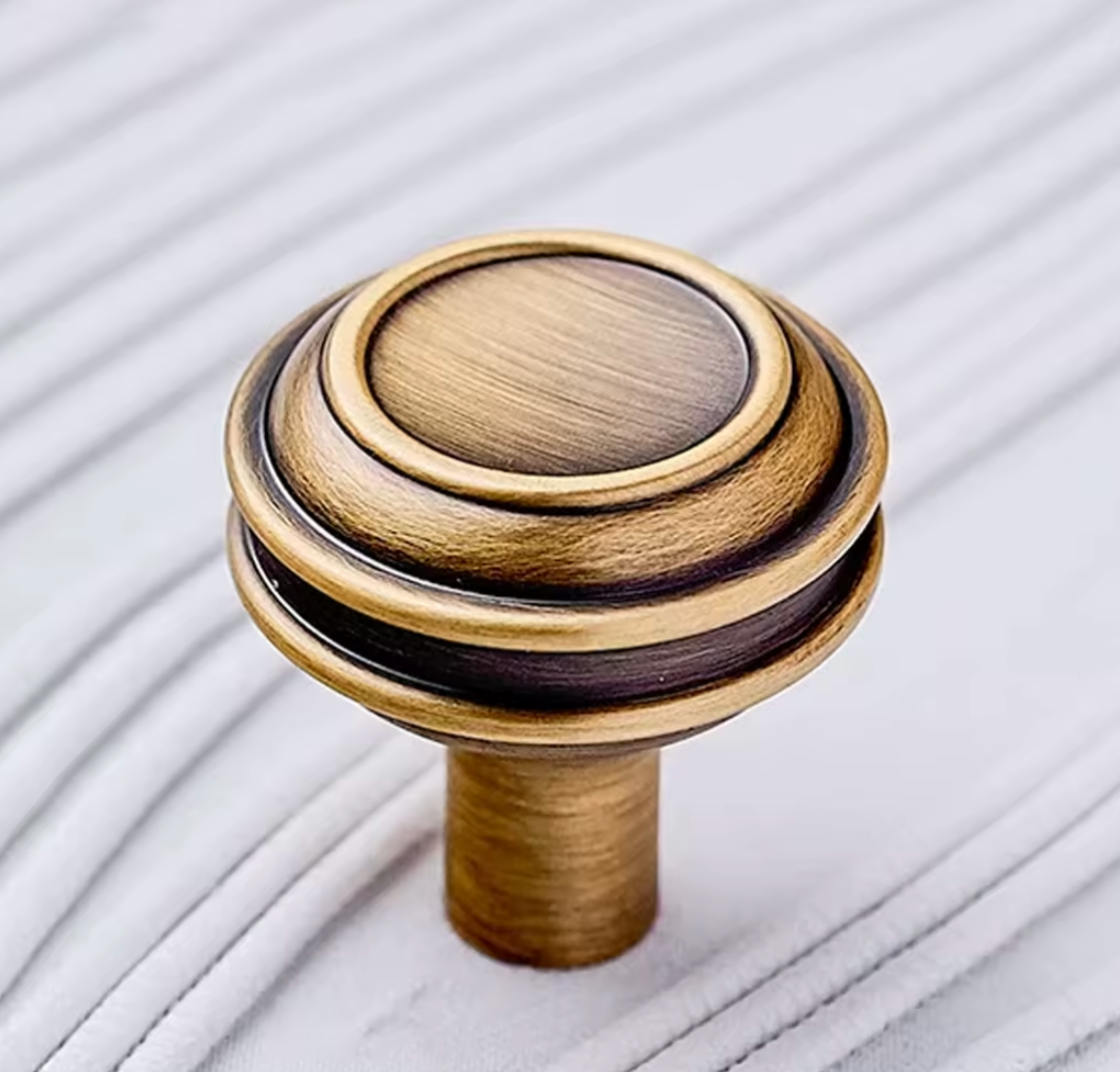 A round wooden cabinet knob with an antique bronze finish and concentric ring detailing, shown on a white textured fabric background.