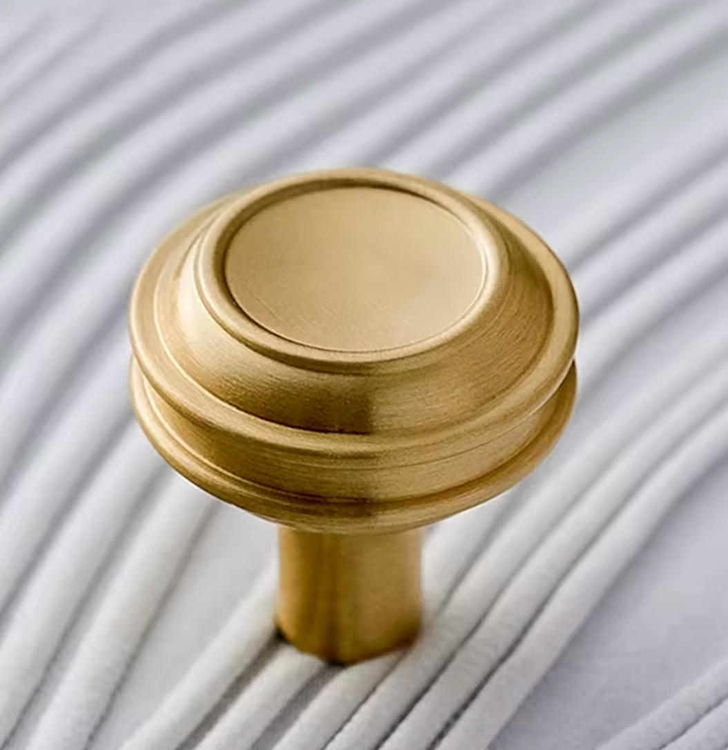 A close-up of a round, brushed brass cabinet knob with a fluted top and cylindrical stem, set against a background of white pleated fabric.