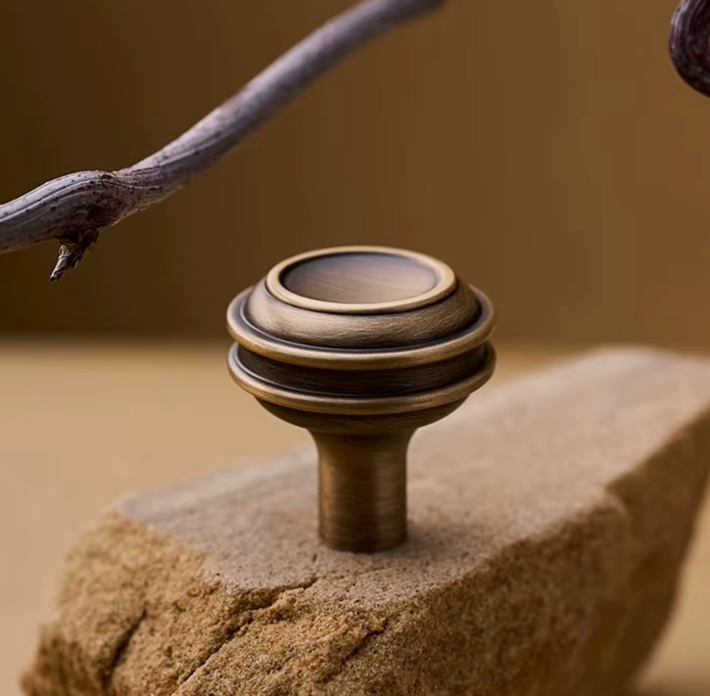 A close-up of a round, tiered cabinet knob with an antique bronze finish, displayed on a textured stone block with a blurred warm brown background.