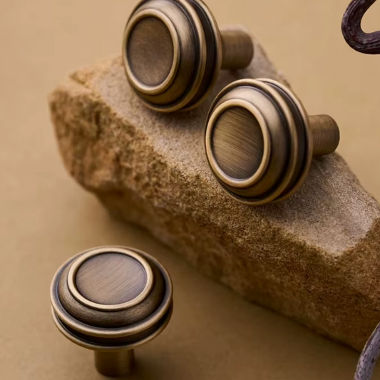 Three round cabinet knobs with a distressed bronze finish and concentric ring detailing, displayed on a textured stone prop against a warm beige background.