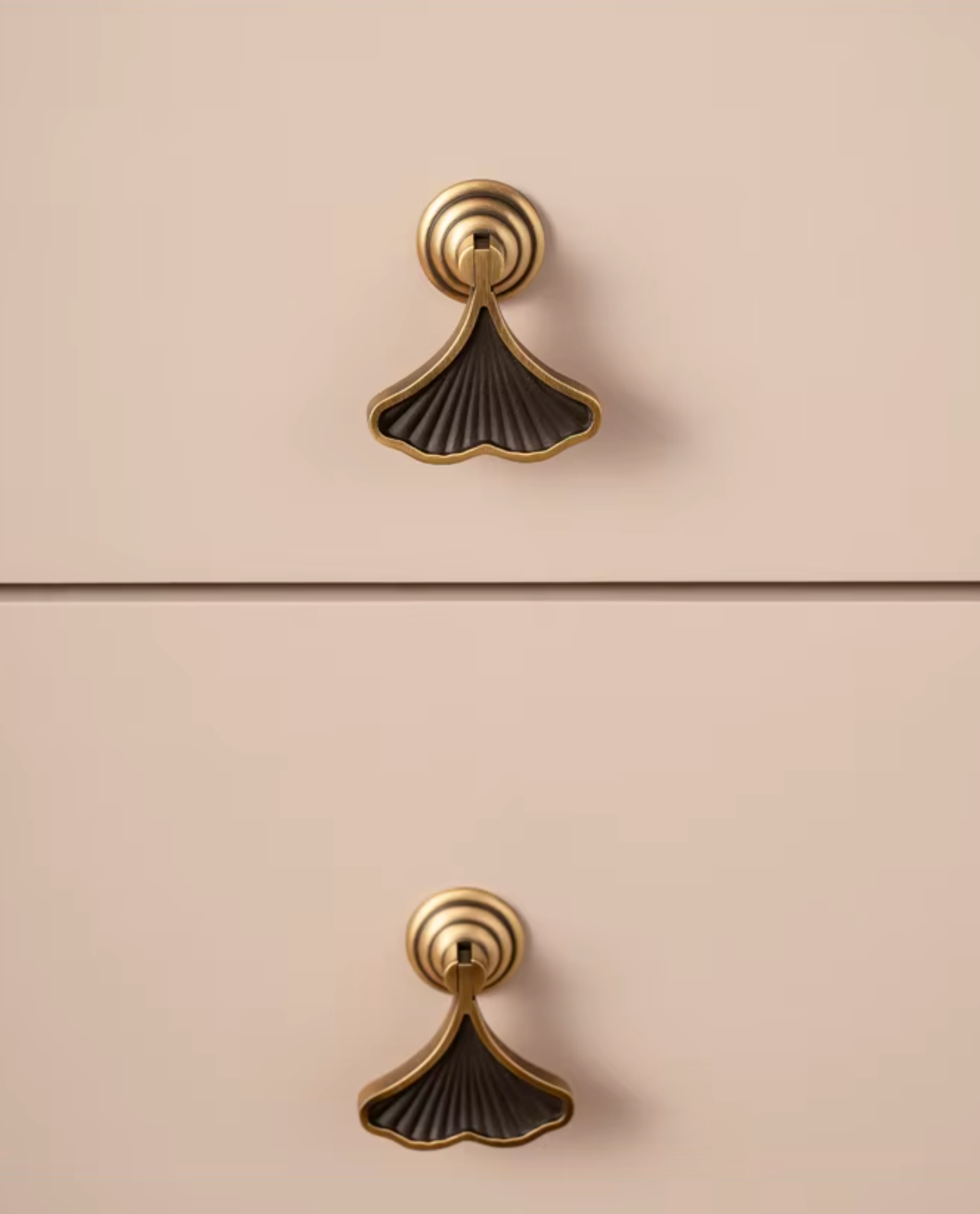 Two matching cabinet drawer pulls shown vertically on a warm beige surface. Each pull has a gold-toned circular top with a fan- or ginkgo-like lower section in a dark bronze/black finish.