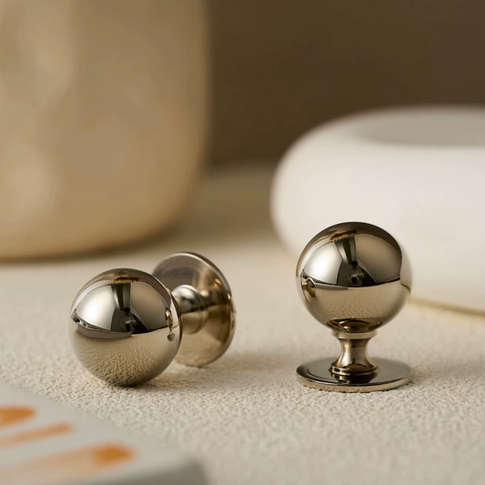 Two polished metallic ball-shaped hardware pieces with round backs, photographed on a light textured surface with a softly blurred neutral background.