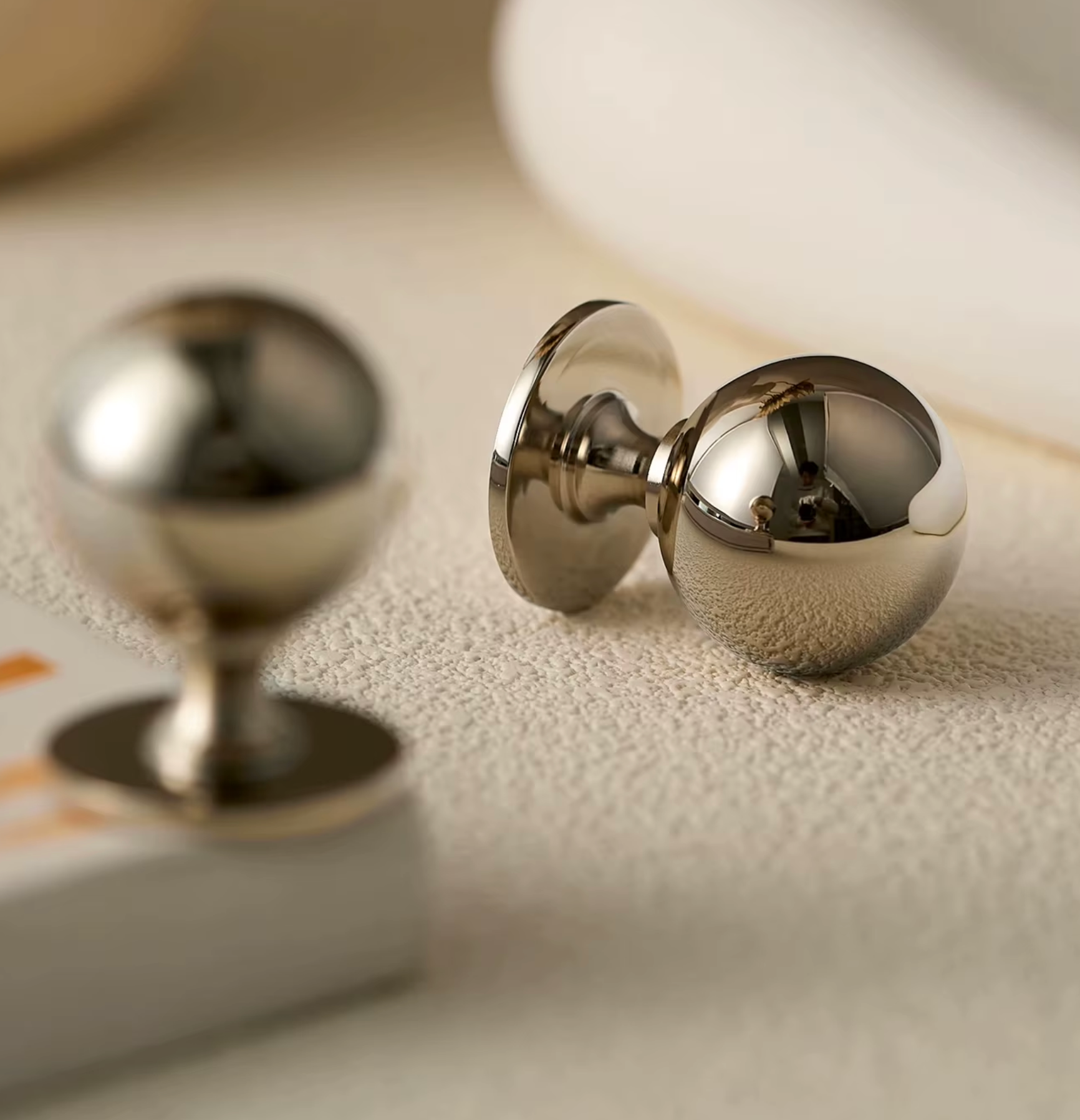 Two polished metallic spherical objects with circular bases, photographed on a soft beige textured surface with shallow depth of field.